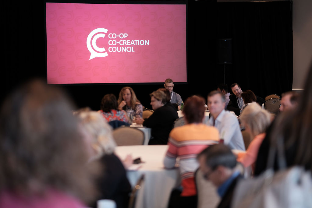 A conference scene at Hyatt, Huntington Beach, with attendees engaged at round tables, and a large screen displaying 'CO-OP CO-CREATION COUNCIL' in the background. business conference hyatt hutington beach photographer nicole caldwell