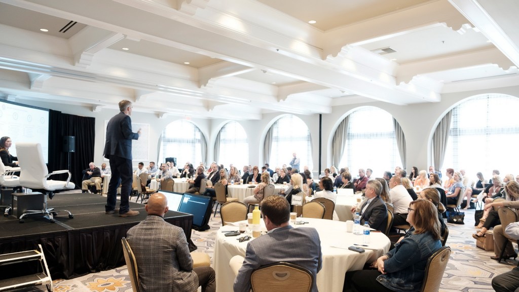 A speaker presenting on stage to an engaged audience at a business conference in a well-lit venue with large windows. business conference hyatt hutington beach photographer nicole caldwell