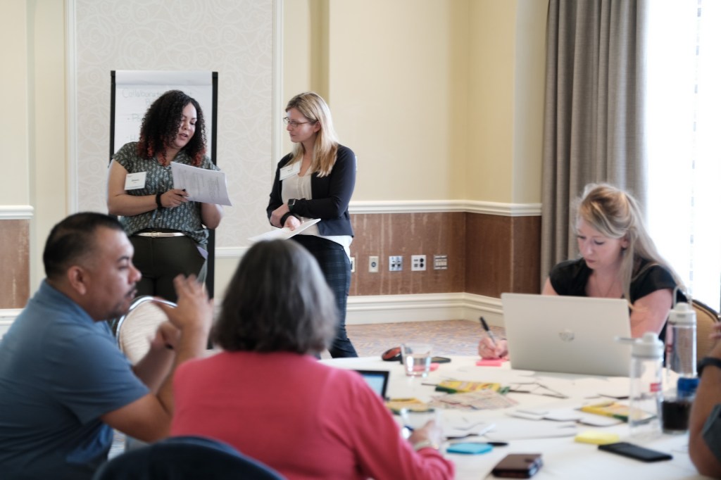 Two women are discussing documents in a conference room during a corporate retreat, while a group of people participates in the discussion at a table filled with papers and laptops. Huntington Beach hyatt orange county business corporate event photographer nicole caldwell