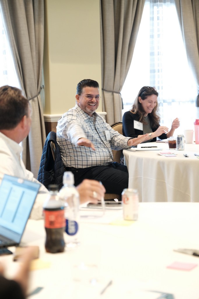 A group of professionals engaged in a discussion at a corporate retreat, with one man smiling and gesturing while a woman sits nearby, both seated at a round table with drinks and laptops. Huntington Beach hyatt orange county business corporate event photographer nicole caldwell