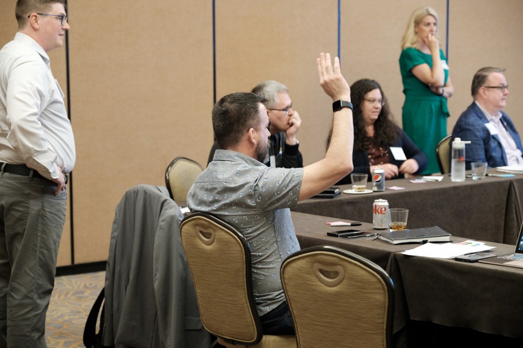 A corporate retreat scene featuring several individuals engaged in a discussion, with one person raising their hand to speak. The setting includes tables with documents and drinks. Huntington Beach hyatt orange county business corporate event photographer nicole caldwell