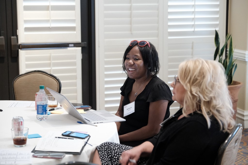 Two women engaged in discussion at a corporate retreat, one smiling and using a laptop, with a table set up with drinks and notes. Huntington Beach hyatt orange county business corporate event photographer nicole caldwell
