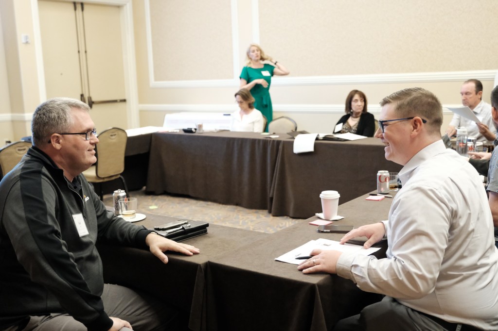 Two men seated at a table engaged in conversation during a corporate retreat, with various attendees in the background. A woman in a green dress stands behind them. Huntington Beach hyatt orange county business corporate event photographer nicole caldwell