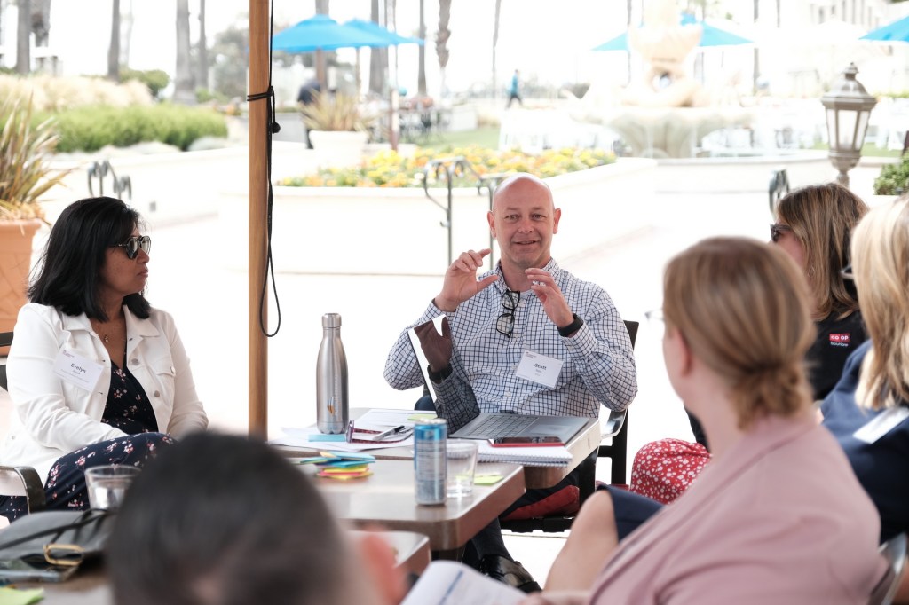 A group of people engaged in discussion during a corporate retreat at Hyatt, Huntington Beach. One man is animatedly speaking while others listen. The setting includes outdoor elements, such as plants and a water feature. Huntington Beach hyatt orange county business corporate event photographer nicole caldwell