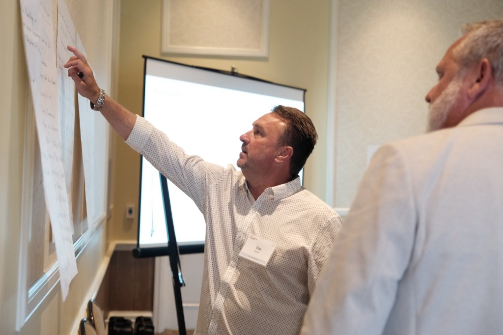 A man pointing at notes on a wall during a corporate retreat, with a presentation screen in the background.