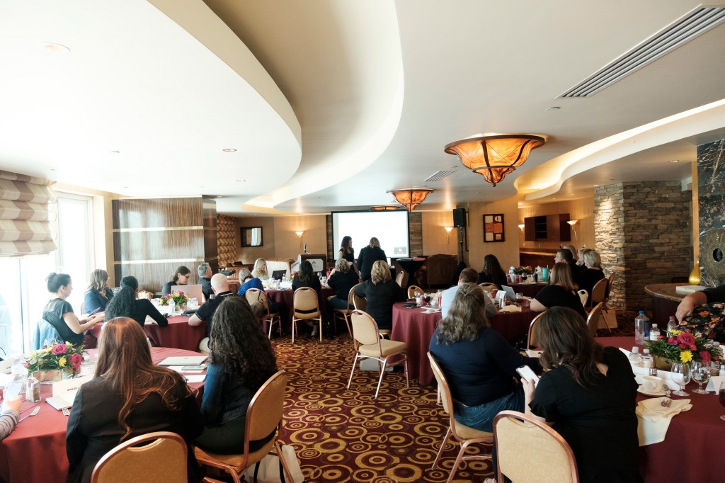 A conference room setting with attendees seated at round tables, watching a presentation on a screen. The room features decorative lighting and floral centerpieces. orange county palm springs headshot business photographer nicole caldwell