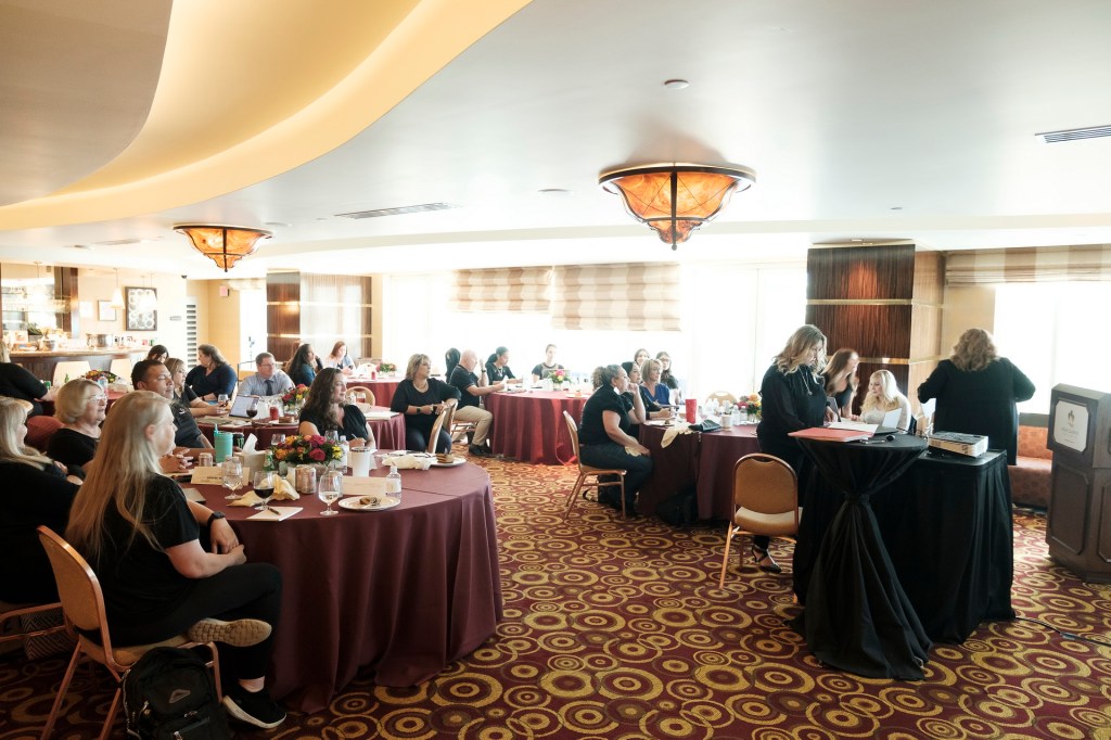 A conference room in Palm Springs, CA, filled with attendees seated at round tables, engaged in discussion. The room features a stylish ceiling design and warm lighting, with refreshments on the tables. orange county palm springs headshot business photographer nicole caldwell