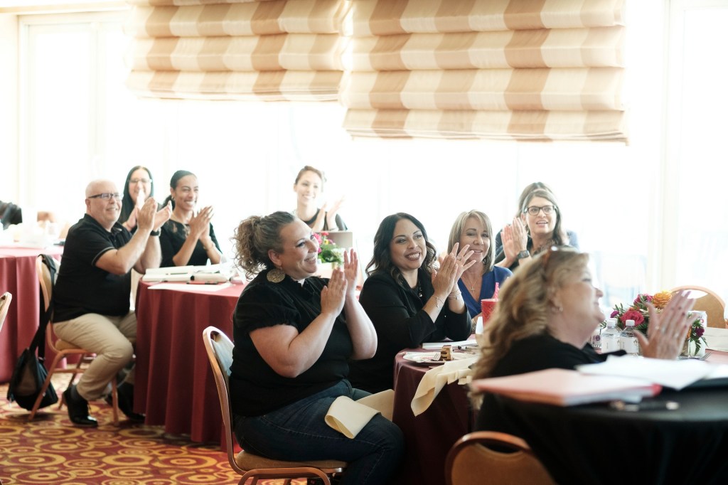 A group of people seated at a table, clapping and smiling during an event, with a bright window in the background and floral decorations on the tables. orange county palm springs headshot business photographer nicole caldwell