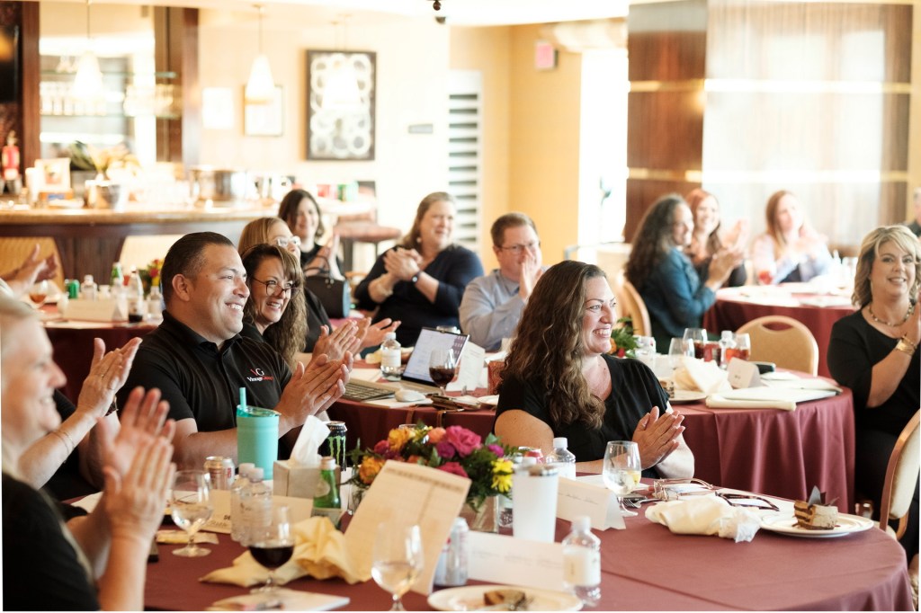 A group of attendees applauding during an event, sitting at tables with drinks and food in a well-lit venue. orange county palm springs headshot business photographer nicole caldwell