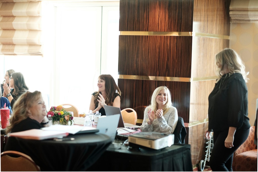 A group of women engaged in conversation and collaboration at a meeting, with one woman standing and smiling while others clap and react positively. The setting includes a large table with laptops and flowers, and large windows allowing natural light. orange county palm springs headshot business photographer nicole caldwell