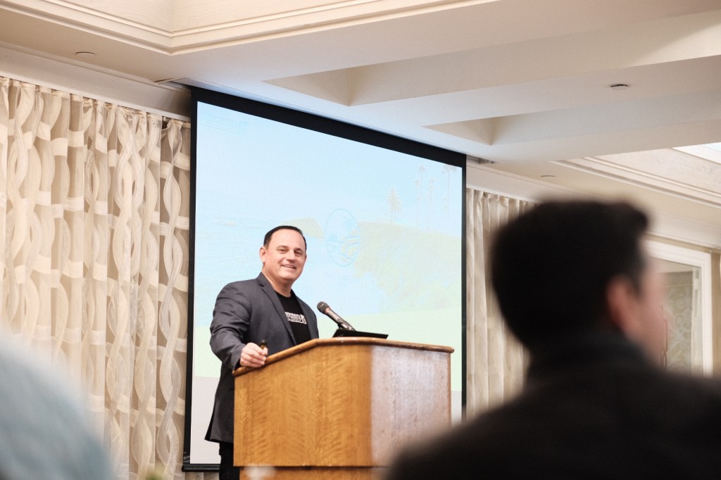 Speaker presenting at a podium during an event in Laguna Beach, CA, with a presentation screen in the background and attendees visible. business conference surf and sand hotel laguna beach photographer nicole caldwell