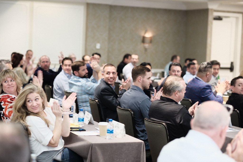 Group of attendees applauding during a welcome reception at a corporate event, seated at tables with water bottles and coffee cups. business conference surf and sand hotel laguna beach photographer nicole caldwell
