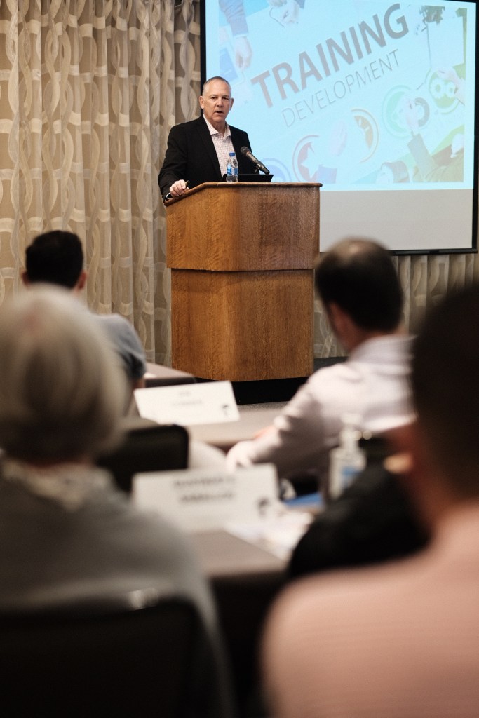 A speaker presenting at a podium during a training development session in a conference room. Attendees are seated in the foreground, with name tags visible. business conference surf and sand hotel laguna beach photographer nicole caldwell
