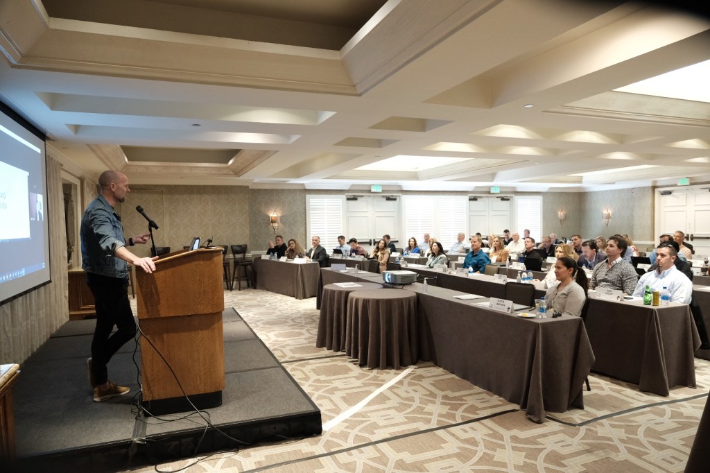 A speaker presenting at a podium in a conference room filled with an audience seated at tables, focusing on the presentation. business conference surf and sand hotel laguna beach photographer nicole caldwell