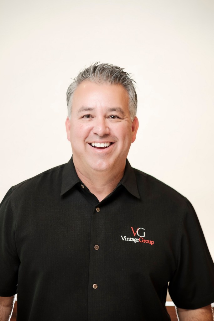 A smiling man wearing a black shirt with 'VintageGroup' logo, standing against a clean white backdrop. orange county palm springs headshot business photographer nicole caldwell