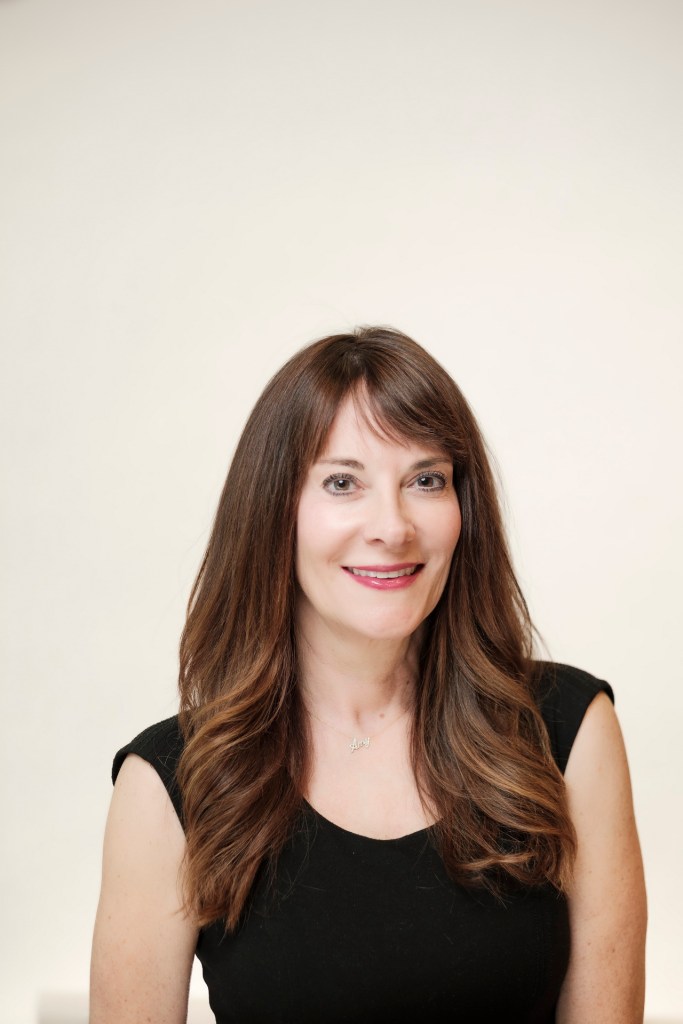 Headshot of a woman with long brown hair, wearing a black top, smiling against a clean white backdrop. orange county palm springs headshot business photographer nicole caldwell