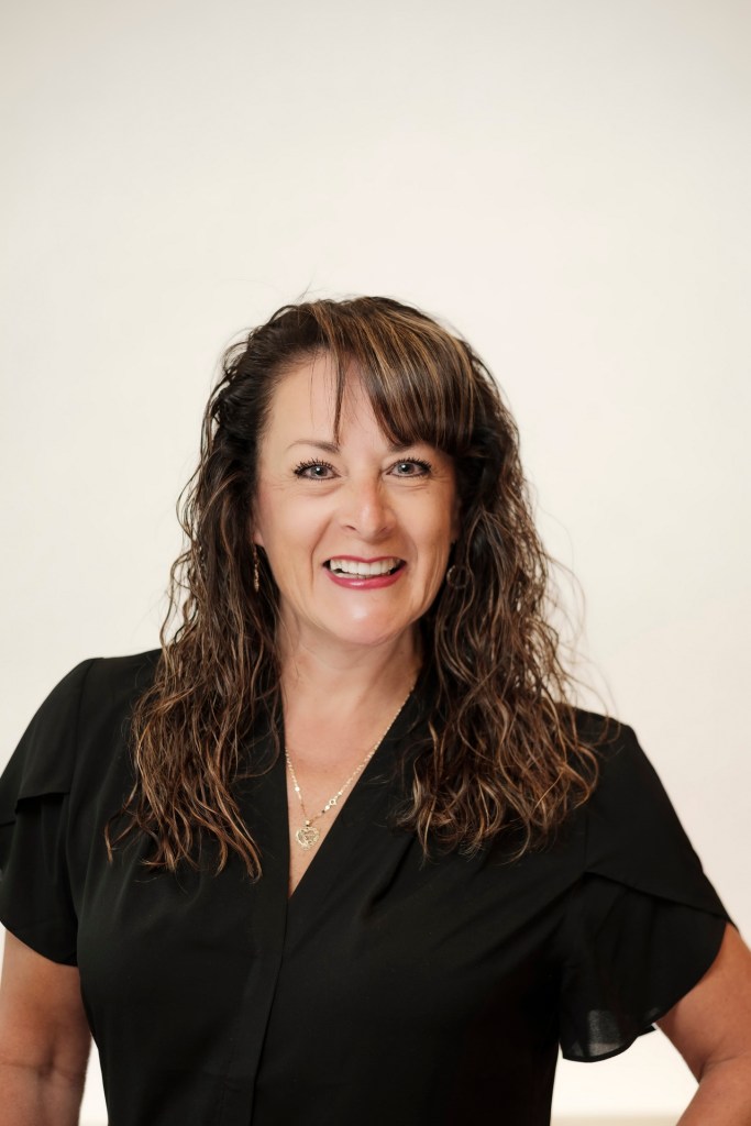 A smiling woman with curly hair wearing a black shirt, standing against a clean white backdrop. orange county palm springs headshot business photographer nicole caldwell