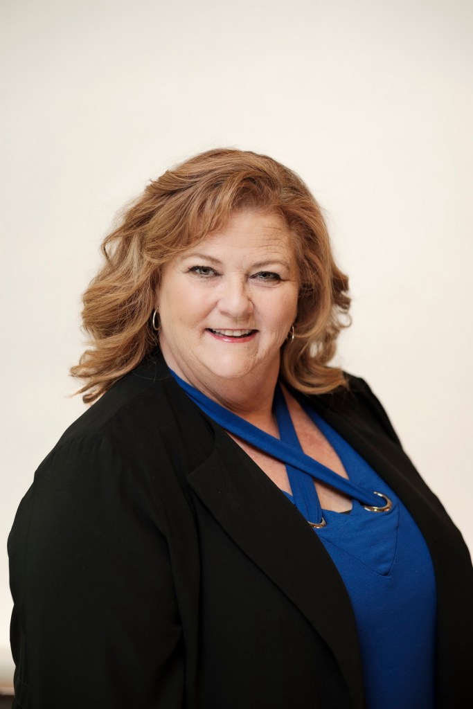 Headshot of a woman with curly brown hair, wearing a black blazer and a blue top, smiling against a clean white backdrop. orange county palm springs headshot business photographer nicole caldwell