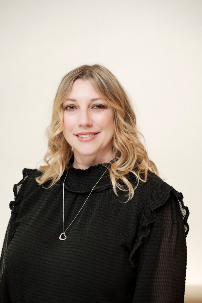 A portrait of a woman with long, wavy blonde hair, wearing a black top with ruffled sleeves and a silver necklace, set against a clean white backdrop. orange county palm springs headshot business photographer nicole caldwell