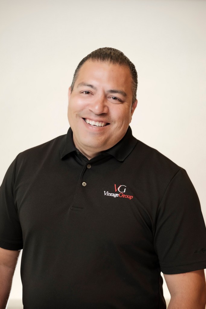 A male subject smiling while wearing a black polo shirt with the Vintage Group logo against a clean white backdrop. orange county palm springs headshot business photographer nicole caldwell