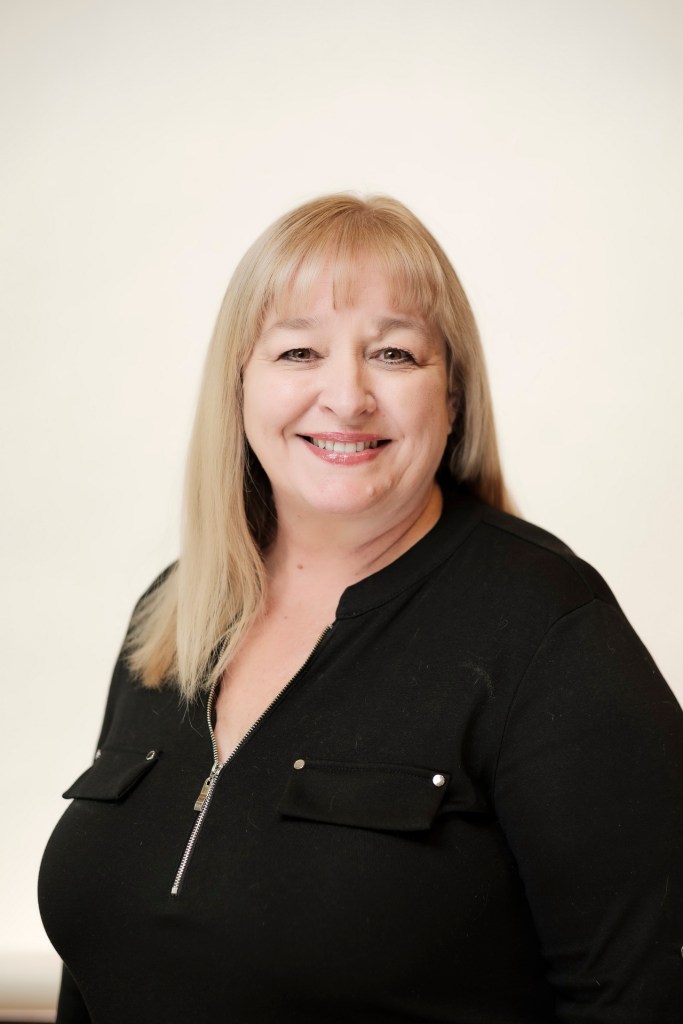 A woman with shoulder-length blonde hair smiling, wearing a black top with a zip neck, posed in front of a clean white backdrop. orange county palm springs headshot business photographer nicole caldwell
