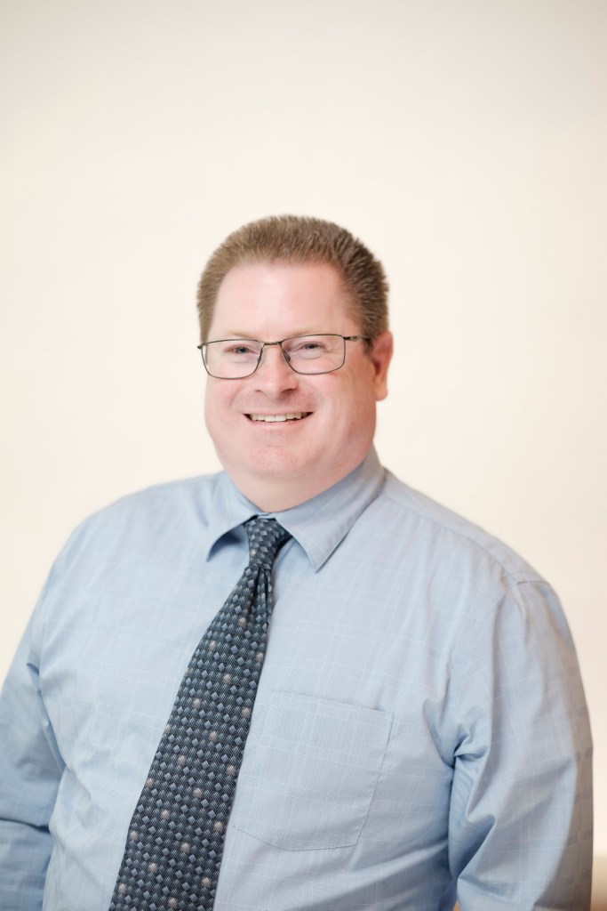 A professional headshot of a smiling man wearing glasses, a light blue dress shirt, and a patterned tie, set against a clean white backdrop. orange county palm springs headshot business photographer nicole caldwell