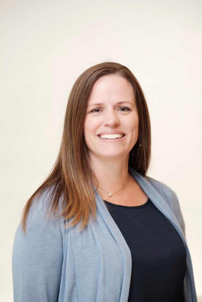 A woman with long brown hair is smiling, wearing a gray cardigan over a black top, against a light-colored backdrop. orange county palm springs headshot business photographer nicole caldwell