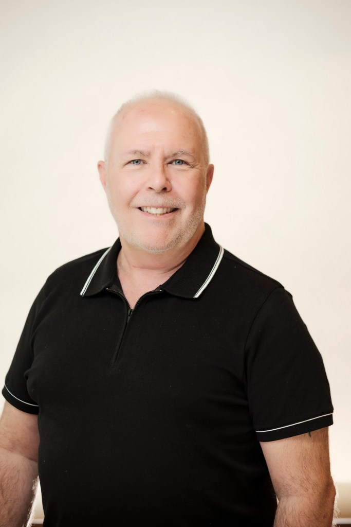 A smiling man with short white hair and a beard is wearing a black polo shirt with white trim, posing against a clean white backdrop. orange county palm springs headshot business photographer nicole caldwell