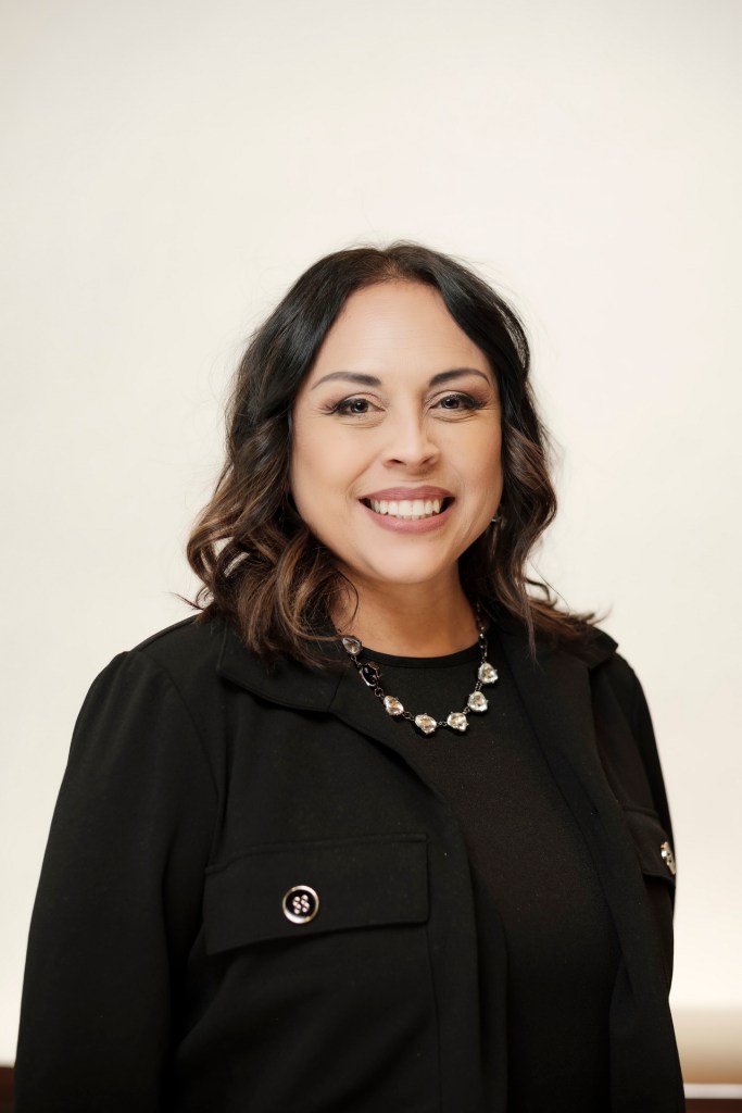 A woman with shoulder-length dark hair smiles warmly. She is wearing a black jacket and a necklace, standing against a clean white backdrop. orange county palm springs headshot business photographer nicole caldwell
