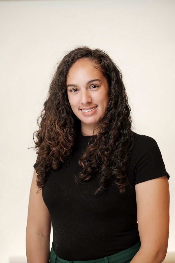 A smiling woman with curly hair is wearing a black shirt in front of a clean white backdrop, posing for a headshot. orange county palm springs headshot business photographer nicole caldwell