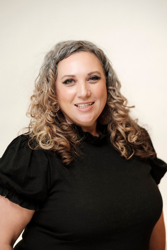 A woman with curly, shoulder-length hair, wearing a black top, smiling against a clean white backdrop. orange county palm springs headshot business photographer nicole caldwell