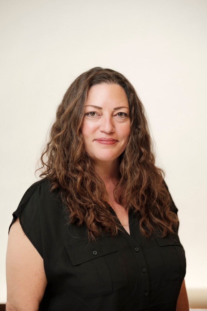 Headshot of a woman with long, curly brown hair, wearing a black shirt, against a clean white backdrop. orange county palm springs headshot business photographer nicole caldwell