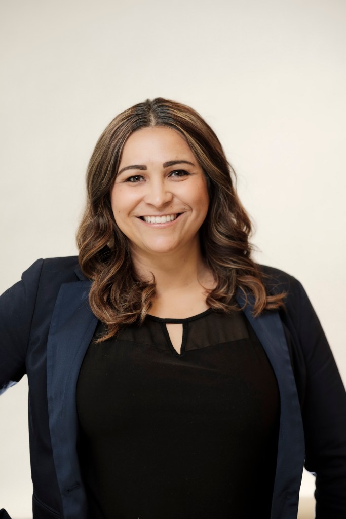 A smiling woman with long, wavy hair wearing a black top and a dark blazer against a clean white backdrop. orange county palm springs headshot business photographer nicole caldwell