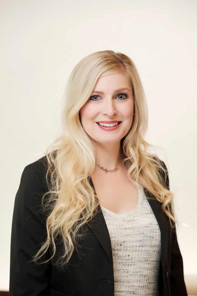 A woman with long blonde hair and a friendly smile, wearing a black blazer over a light top, poses against a soft white backdrop. orange county palm springs headshot business photographer nicole caldwell