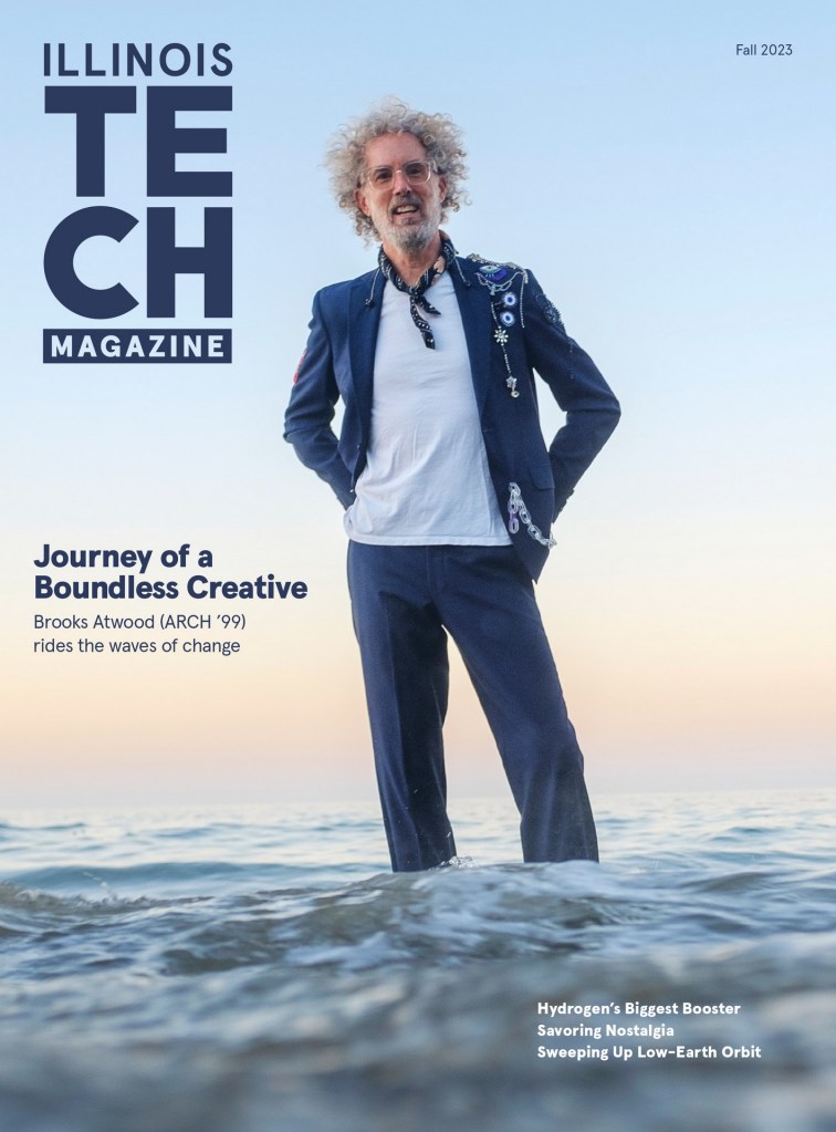 A man with curly hair and glasses stands in shallow water at sunset, wearing a stylish outfit with a blue blazer and a white shirt, in Laguna Beach, CA.executive headshots portraits orange county photographer nicole caldwell