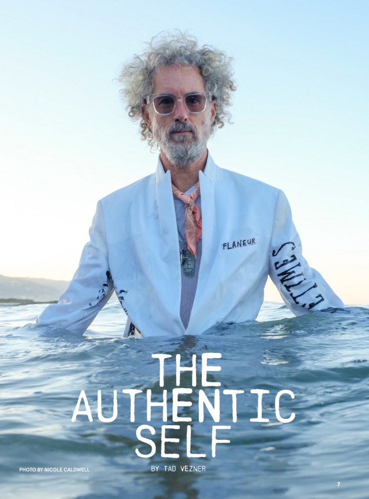 A person with curly hair and glasses stands in the ocean, wearing a white jacket with 'FLANEUR' printed on it. The background features a clear sky and distant shoreline. Laguna Beach, CA executive headshots portraits orange county photographer nicole caldwell
