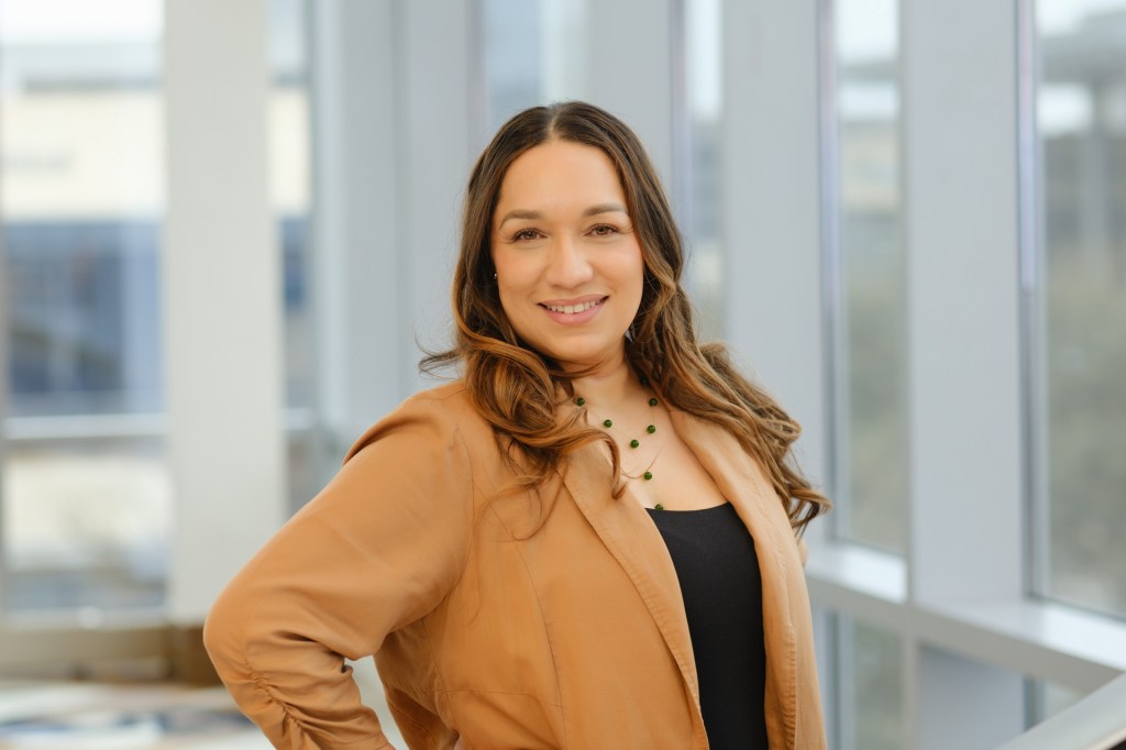 A smiling woman with long, wavy hair poses confidently in a well-lit setting with large windows in the background. She is wearing a tan jacket and a black top, accessorized with a green necklace. omni dallas headshot photographer nicole caldwell