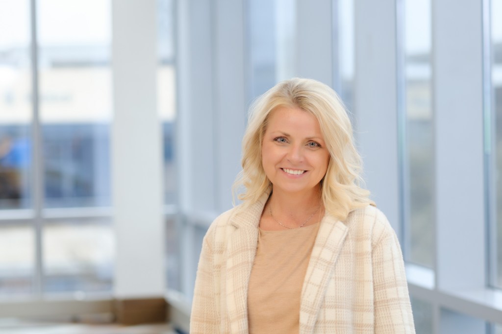 A smiling woman with blonde hair wearing a beige outfit and a light plaid cardigan stands in a well-lit environment with large windows in the background. omni dallas headshot photographer nicole caldwell