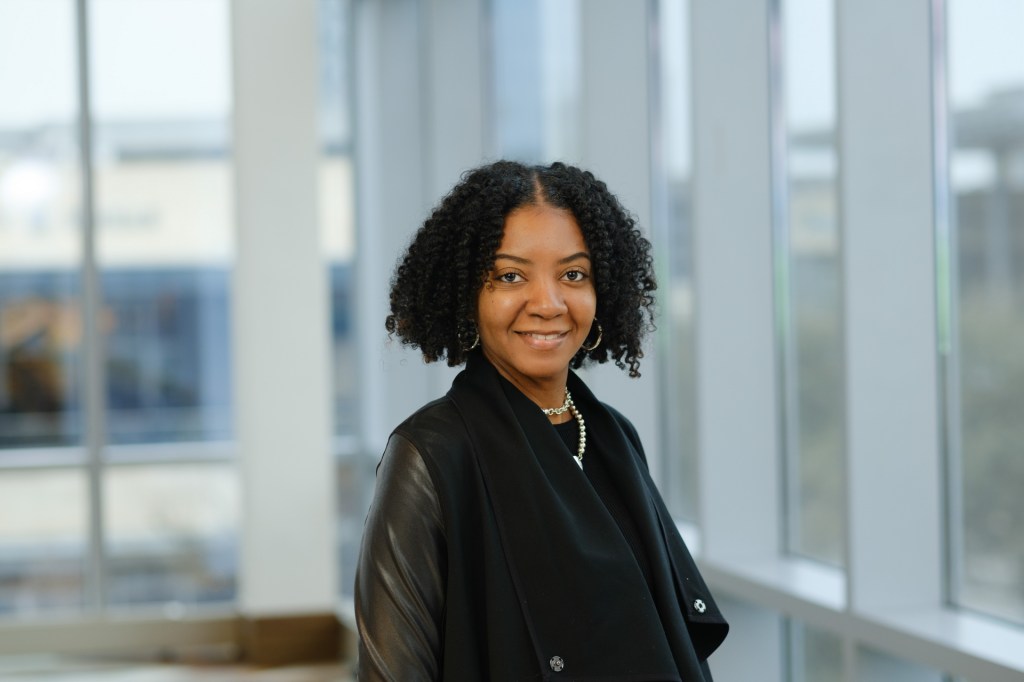 Portrait of a woman with curly hair, wearing a black jacket, smiling in front of large windows in a contemporary setting. omni dallas headshot photographer nicole caldwell