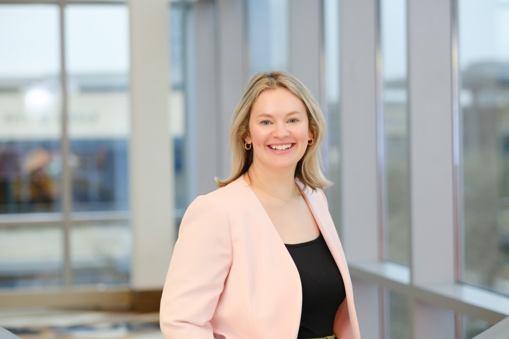 A smiling woman in a pink blazer stands by a window, with a blurred background of a hotel lobby. omni dallas headshot photographer nicole caldwell