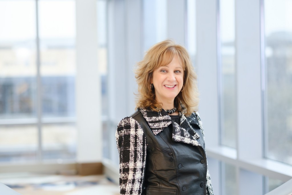A smiling woman with wavy hair stands in a bright, modern indoor space, wearing a black and white jacket. omni dallas headshot photographer nicole caldwell