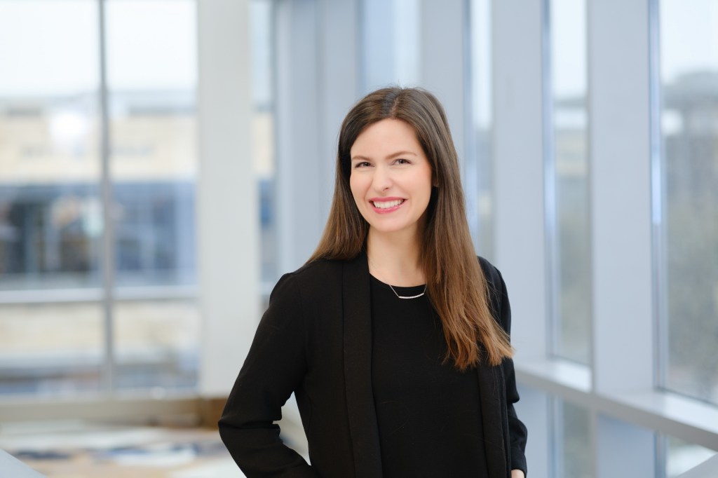 A professional woman smiling, wearing a black outfit, posing indoors with large windows in the background. omni dallas headshot photographer nicole caldwell