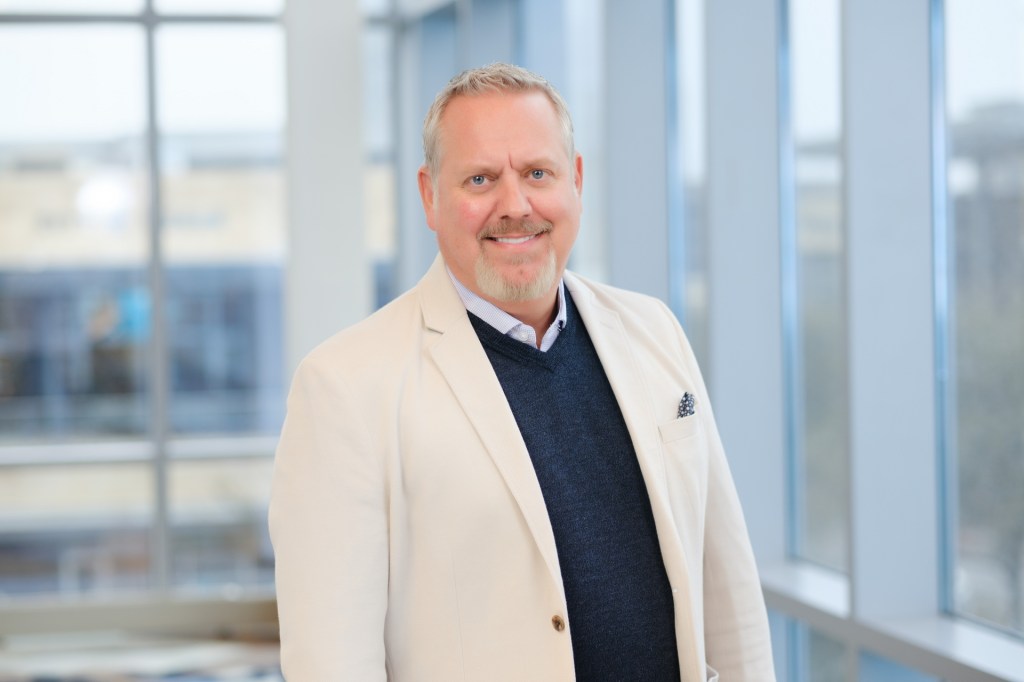 A professional headshot of a smiling man in a light-colored blazer and navy sweater, standing near large windows in a modern office environment. omni dallas headshot photographer nicole caldwell