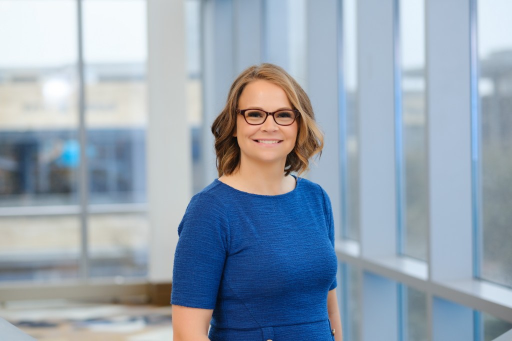 A smiling woman with shoulder-length hair wearing glasses and a blue dress stands in front of large windows, showcasing a bright and professional background. omni dallas headshot photographer nicole caldwell