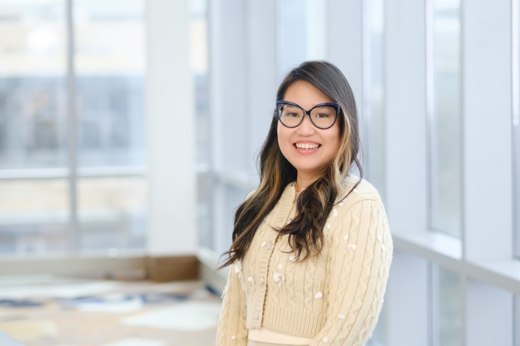 A smiling individual wearing a cream-colored sweater stands in front of large windows, showcasing a bright and airy environment, during a seminar in Dallas, Texas. omni dallas headshot photographer nicole caldwell