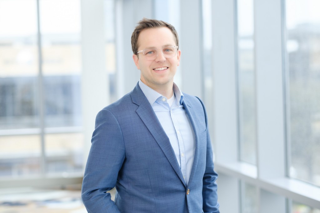 A professional man in a blue suit and glasses smiles while standing near a large window with natural light illuminating the space. omni dallas headshot photographer nicole caldwell