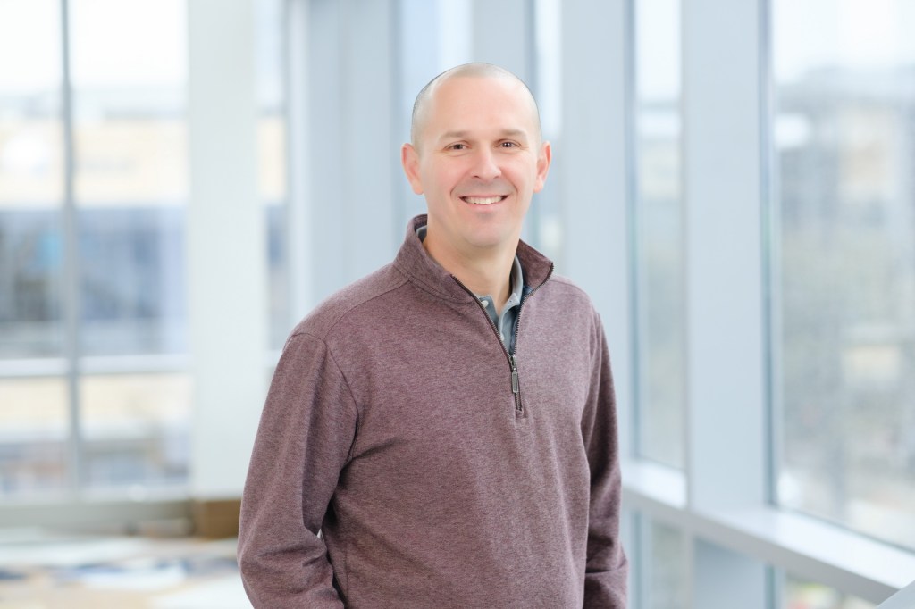 A smiling man in a burgundy sweater stands in front of large windows, with a blurred cityscape visible outside. omni dallas headshot photographer nicole caldwell
