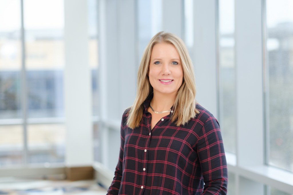 Professional portrait of a smiling woman with long blonde hair, wearing a plaid shirt, standing in a well-lit interior with large windows. omni dallas headshot photographer nicole caldwell