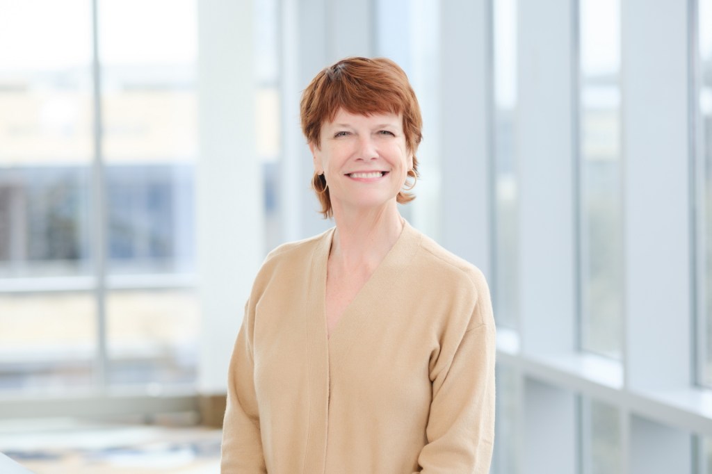 A smiling woman with short red hair wearing a beige sweater stands in front of large windows, showcasing a bright and airy environment. omni dallas headshot photographer nicole caldwell