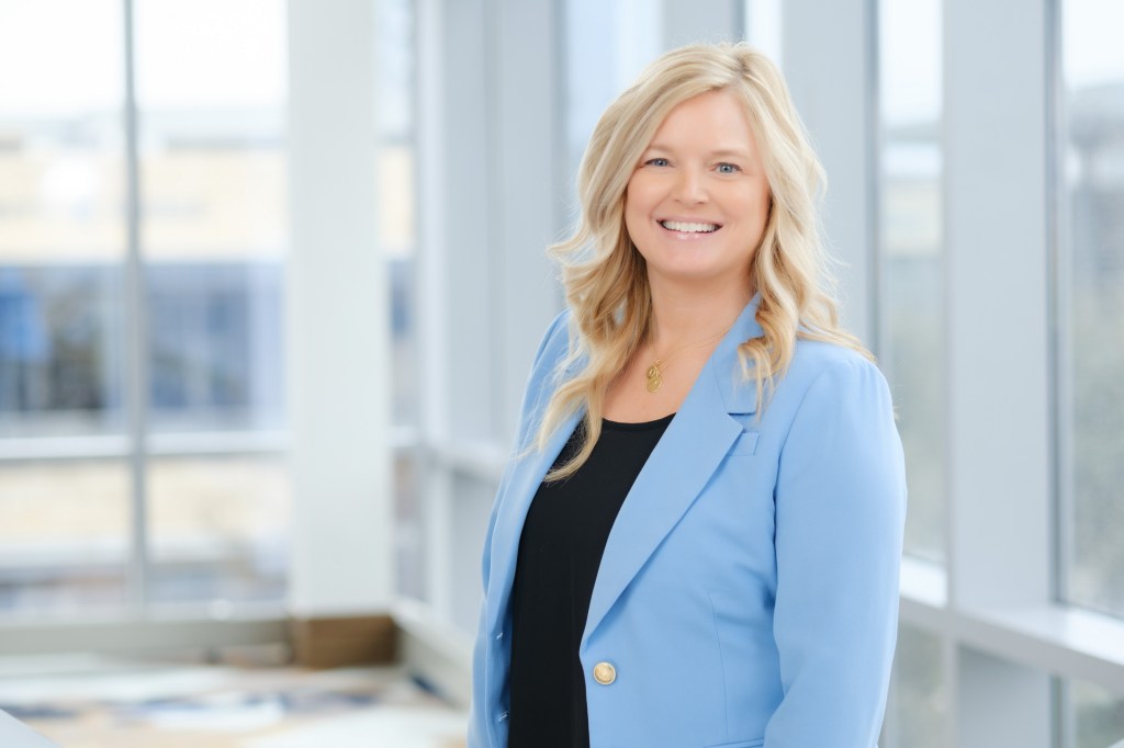 A woman smiling in a light blue blazer stands in front of large windows in a conference setting. omni dallas headshot photographer nicole caldwell